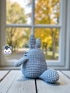 Blue and white crochet Totoro amigurumi on a wooden table with a blurry autumn forest background.