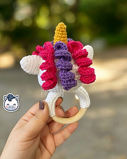 Crochet amigurumi pattern of a baby unicorn rattle with a golden horn, pink and purple mane on a wooden ring.