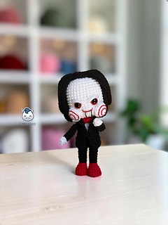 Handmade Billy the Puppet amigurumi inspired by Saw, a horror crochet doll with black suit, red bow tie, red eyes, spiral cheeks and black hair, perfect for Halloween crochet collections.