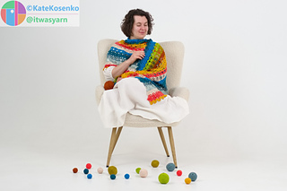 Woman wrapped in modular crochet shawl, sitting in chair with yarn balls, cozy handmade scene