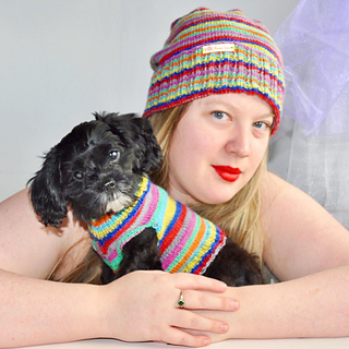 A woman with blonde hair and ruby lipstick wearing a striped hat with her arms around a little black adorable dog who is wearing a matching striped sweater.