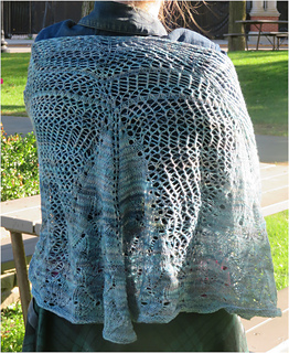 Image Description: A person wearing a pale blue, half-circle knit shawl. The top edge of the shawl is a mesh stitch, which transitions into stocking stitch, then into a lace pattern stitch near the bottom of the shawl.
