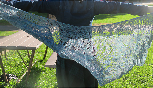 Image Description: a pale blue lace shawl held out behind a person.  It's a half-circle shawl knit in a starburst mesh pattern with a lace pattern stitch at the bottom edge. 