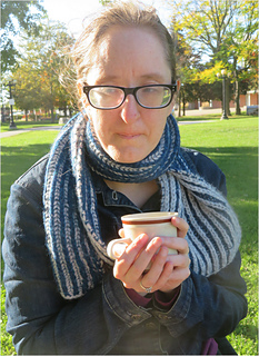 Image Description: A woman wearing a bulky-knit reversible scarf and holding a mug.  The scarf is knit in two-colour fisherman's ribbing, forming vertical stripes in two colours, off white and dark blue.