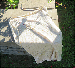 Image Description: A cream skirt laying flat on the edge of a garden bed. The skirt is a wrap skirt, knit in stocking stitch with I-cord ties.  5 buttons fasten the front edge of the skirt, and the lower hem is edged with eyelets