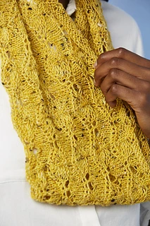[Image Description: A close up of a knit lace cowl, worked in a scalloped lace pattern.  The yarn is fingering-weight yarn in a mustard yellow with flecks of black.  Someone is holding one edge of the cowl in their hand.]