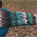 [Image Description: A woman holding a scarf across her shoulder and back with an outstretched arm. The scarf is knit in striping worsted-yarn, and shows a diamond lace pattern.]
