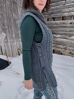A female is standing side view in front of a barn wall wearing a blue cabled vest and a brown leather belt-pouch. Showing detail of side buttons on vest.