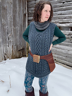 A female is standing with hands on hips facing the camera in front of a barn wall wearing a blue cabled vest and a brown leather belt-pouch.