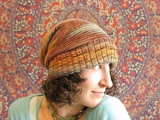 Brunette woman with curly hair wearing a multi-coloured slouchy knit hat with the ribbed brim folded up and a botanical (tapestry) background.