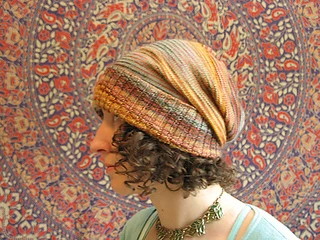 Brunette woman with curly hair wearing a multi-coloured slouchy knit hat with the ribbed brim folded up and a botanical (tapestry) background.