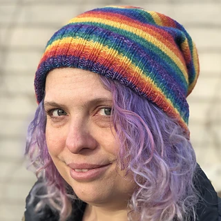 A woman looking at the camera with curly purple hair and wearing a slouchy hat with rainbow stripes.