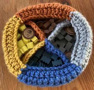 A crocheted bag with four sections holding four different board game tokens. The sections are made with brown, grey, blue, and gold yarn, to match the tokens. 