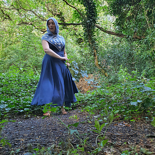 Woman wearing blue and grey crocheted fancy granny square hood in woodland setting (hood up/front view)