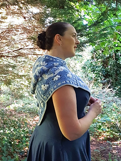 Woman wearing blue and grey crocheted fancy granny square hood in woodland setting (hood down/side view)