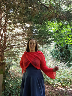 Smiling woman in coral crochet lace wrap cardigan tied  at each shoulder stands in a forest, one arm outstretched