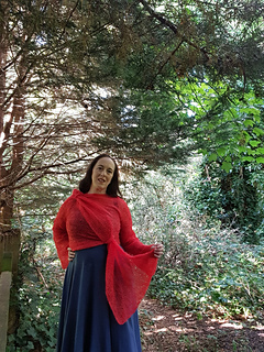 Smiling woman in coral crochet lace wrap cardigan tied asymmetrically at hip and shoulder stands in a forest, hand on one hip