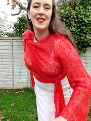 Smiling woman in coral crochet lace wrap cardigan tied asymmetrically at hip and shoulder stands in a garden leaning forwards