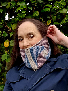 Woman in front of green foliage, wearing a blue coat and a knitted spiral striped cowl held up over the lower half of her face.