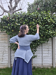 Woman in a blue crocheted tunic poses facing away reaching to a bush in a garden with green bushes and bare tree. Wooden fence in the background. Calm expression.