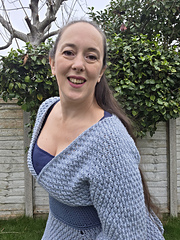 Woman in a blue crocheted tunic poses leaning forward in a garden with green bushes and bare tree. Wooden fence in the background. Happy expression.