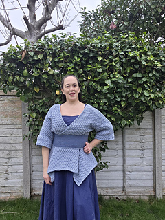 Woman in a blue crocheted tunic poses looking ahead in a garden with green bushes and bare tree. Wooden fence in the background. Calm expression.