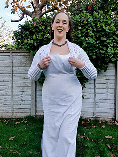 Woman in a white knitted shrug smiles outdoors. Greenery and a wooden fence are in the background on a sunny day.