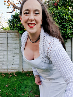 Woman in a white knitted shrug smiles outdoors. Greenery and a wooden fence are in the background on a sunny day.