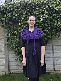 Woman in a black dress with a purple variegated hairpin crocheted shawl stands in a garden. Greenery and a fence are in the background.