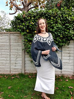 Woman smiling in a garden wearing a blue and white poncho. Wooden fence and leafless trees in the background under a cloudy sky.