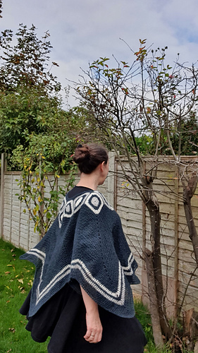 Woman smiling in a garden wearing a blue and white poncho. Wooden fence and leafless trees in the background under a cloudy sky.