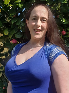 Woman wearing a blue and white knitted shrug stands in a sunny garden, with a leafy background. She looks ahead, smiling.