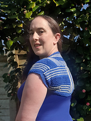 Woman wearing a blue and white knitted shrug stands in a sunny garden, with a leafy background. She looks over her shoulder, smiling.
