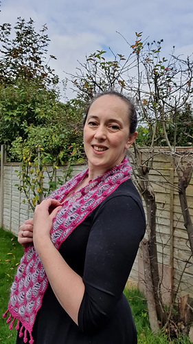 Smiling woman in a black dress with a pink and light blue crochet scarf stands in a garden with green hedges and a tree.