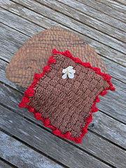 Brown basketweave knitted square with red bobble border and a white flower on top, placed on a wooden stand.