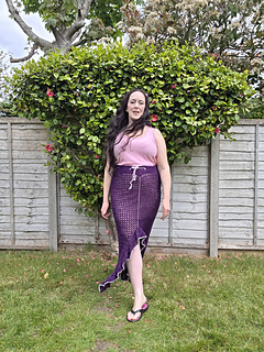 A woman in a pink top and long purple crocheted ruffle edged split skirt stands on grass, backed by a garden fence and lush green hedge with flowers. Overcast sky above.