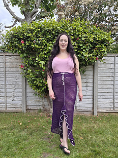 A woman in a pink top and long purple crocheted ruffle edged split skirt stands on grass, backed by a garden fence and lush green hedge with flowers. Overcast sky above.
