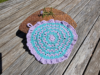 Crocheted wash cloth in purple and teal on wooden table, with rosemary and small purple flower atop a brown woven mat.