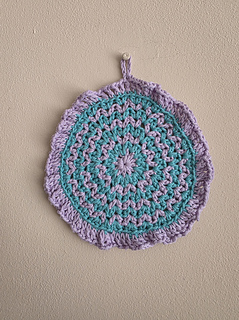 Crocheted round teal and lavender wash cloth with a hanging loop against a beige wall, showcasing an intricate pattern.