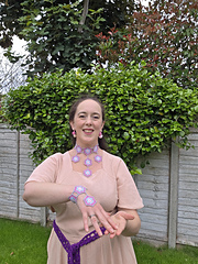 Smiling woman in a garden, wearing a pink dress and crocheted hexagon jewellery, posing before a leafy hedge.