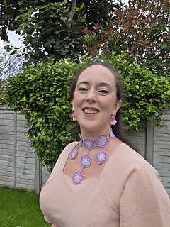 Smiling woman in a garden, wearing a pink dress and crocheted hexagon jewellery, posing before a leafy hedge.