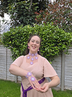 woman at a table, wearing a brown top and crocheted hexagon jewellery, posing with wrist and neck visible