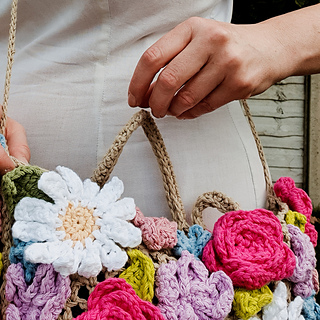 Smiling woman in a white dress holds a crocheted flower-covered bag in a leafy garden beside a wooden fence.
