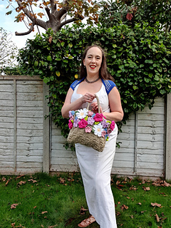 Smiling woman in a white dress holds a crocheted flower-covered bag in a leafy garden beside a wooden fence.