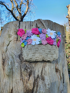Smiling woman in a white dress holds a crocheted flower-covered bag in a leafy garden beside a wooden fence.