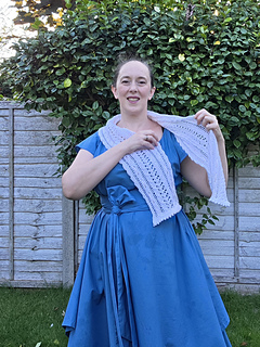 Woman in a blue dress smiles while putting on a white lace knitted scarf with pink ribbons woven through in a leafy garden.