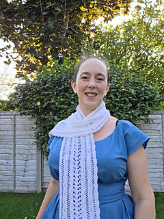 Woman in a blue dress smiles while wearing white lace knitted scarf with pink ribbons woven through in a leafy garden.