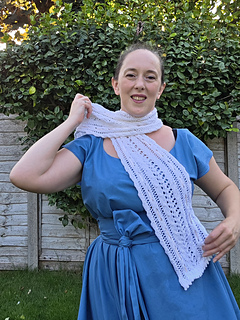 Woman in a blue dress smiles while putting on awhite lace knitted scarf with pink ribbons woven through in a leafy garden.