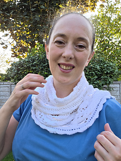 Woman in a blue dress smiles while gesturing to her white lace knitted scarf with pink ribbons woven through in a leafy garden.