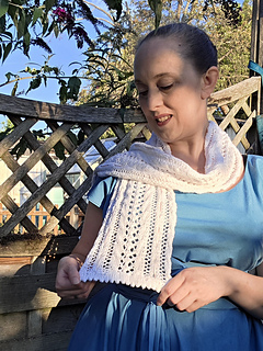Woman in a blue dress smiles while holding up a white lace knitted scarf with pink ribbons woven through in a leafy garden.